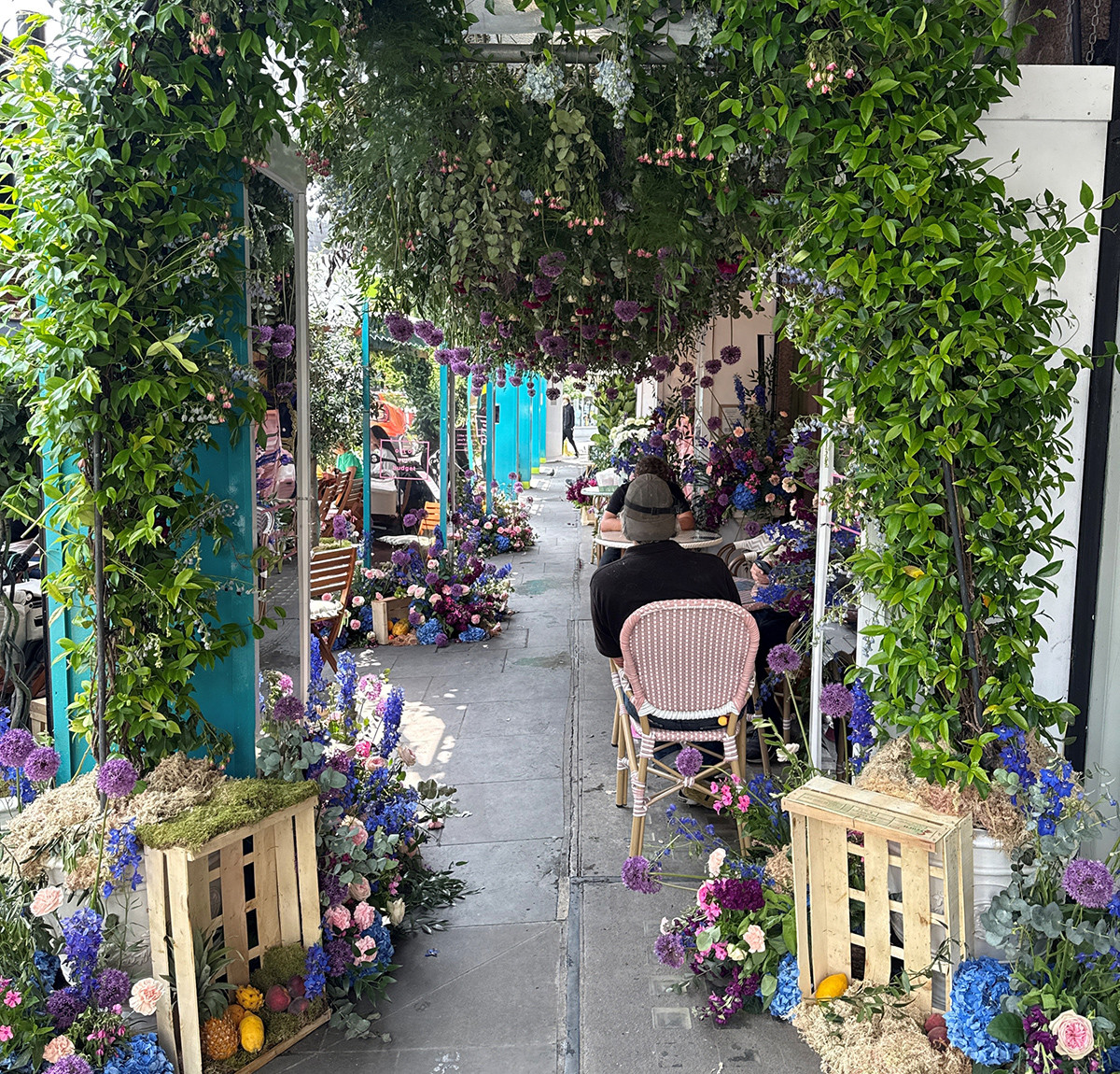A flower-filled sidewalk café with lush greenery overhead and people seated at small tables along a decorated walkway.