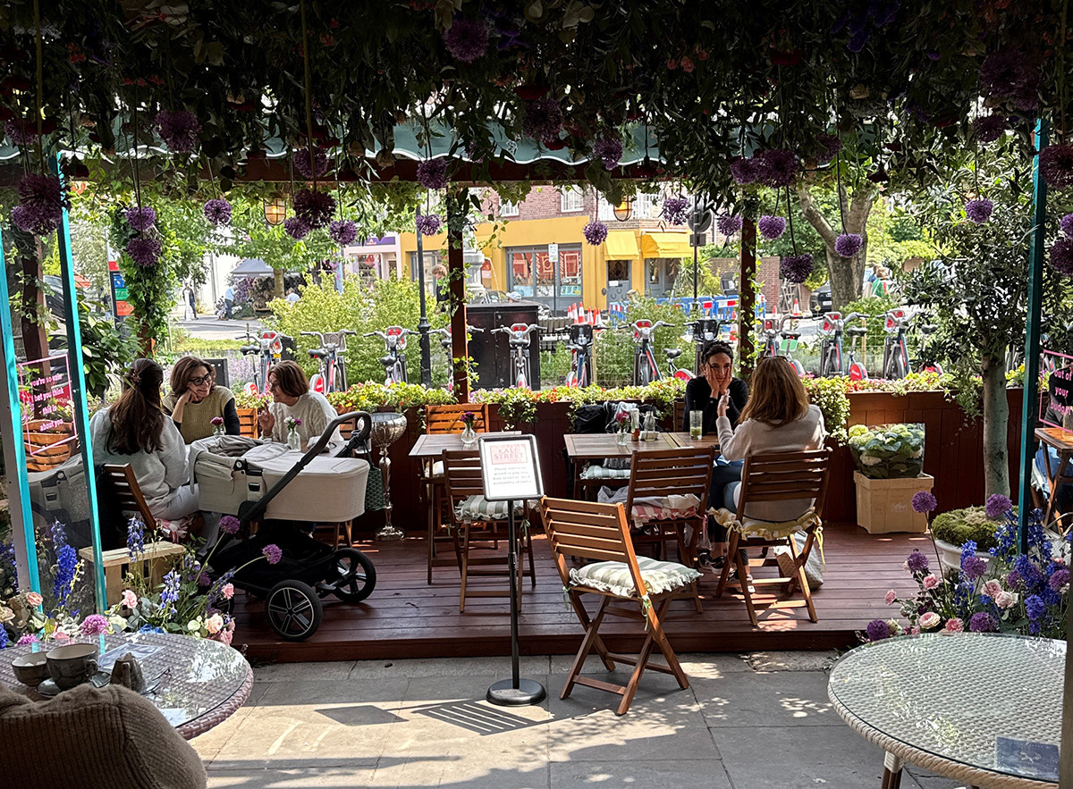 A flower-filled sidewalk café with hanging blooms overhead and people seated at small tables along a decorated patio.