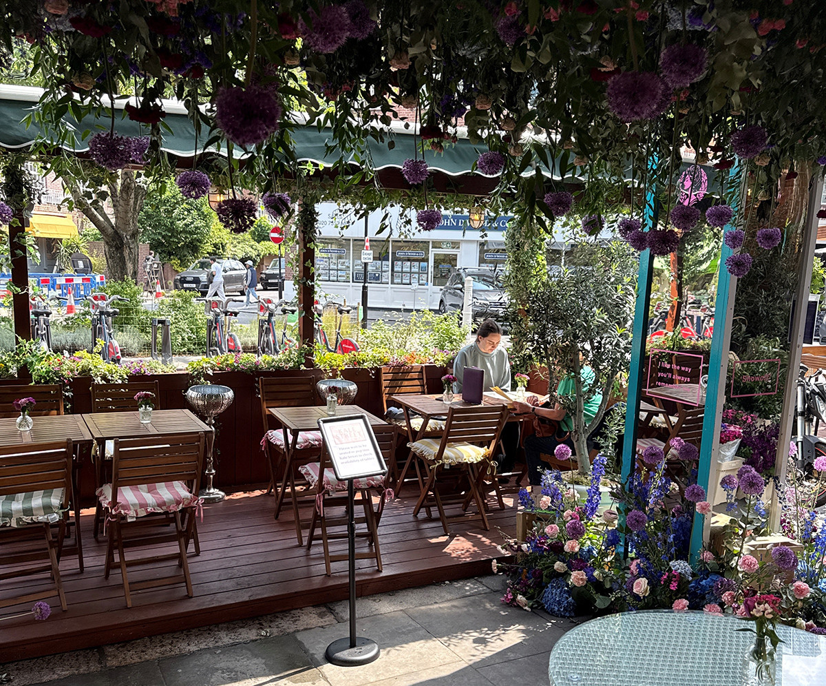 A flower-filled sidewalk café with hanging blooms overhead and people seated at small tables along a decorated patio.